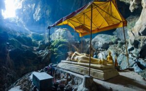 Statue de Bouddha couché sous un auvent dans une grotte sacrée au Laos, baignée de lumière naturelle filtrant par l’entrée, entourée de formations rocheuses et d’objets d’offrandes