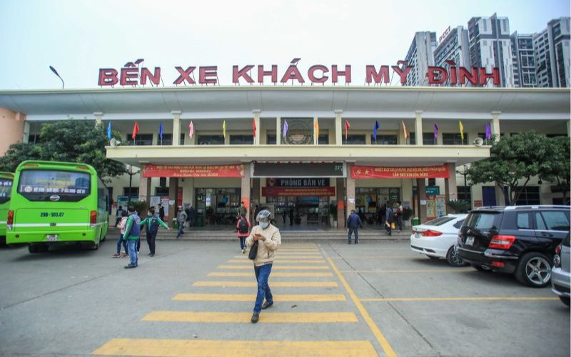 Entrée principale de la gare routière de My Dinh à Hanoï, avec des bus verts stationnés, des voyageurs et le panneau « Bến xe khách Mỹ Đình » visible en haut du bâtiment