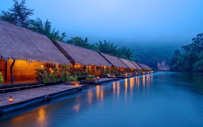 Hôtel flottant traditionnel en bambou éclairé au bord de la rivière Kwai, entouré de jungle luxuriante et de brume matinale à Kanchanaburi