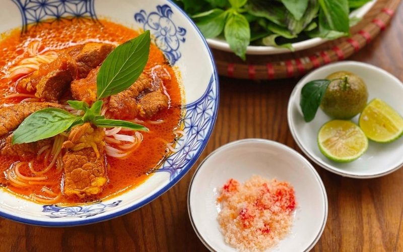 Un bol de hủ tiếu cà ri Vĩnh Châu aux nouilles de riz, servi avec du porc mijoté dans une sauce au curry rouge onctueuse, décoré de basilic frais. À côté, des quartiers de citron vert et un petit plat de sel au piment pour relever les saveurs