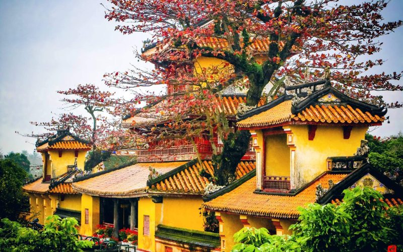 Vue pittoresque de bâtiments traditionnels aux toits en tuiles orangées dans la Cité impériale de Hué, entourés d’arbres aux feuilles rouges d’automne, créant une atmosphère paisible et poétique