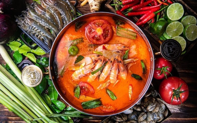 Bol de soupe thaïlandaise tom yum aux crevettes, entouré d’ingrédients frais comme citronnelle, piments rouges, citrons verts, tomates et herbes aromatiques