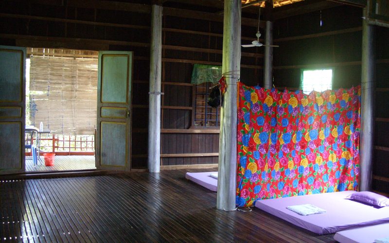 Intérieur simple d’une maison traditionnelle cambodgienne en bois, avec des matelas posés au sol et une cloison colorée séparant l’espace de couchage