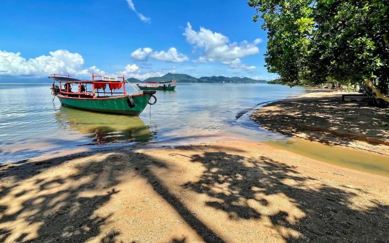 Plage paisible de l’île aux Lapins (Koh Tonsay) au Cambodge, avec des bateaux traditionnels amarrés sur une mer calme, entourée de collines verdoyantes sous un ciel bleu éclatant