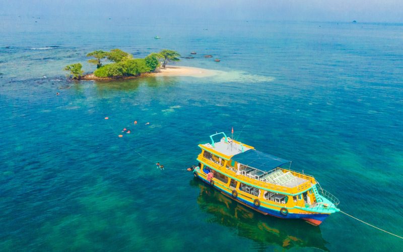 Un bateau de croisière coloré ancré dans une mer turquoise près d’un petit îlot verdoyant, avec plusieurs personnes nageant et faisant du snorkeling dans les eaux claires de Phu Quoc