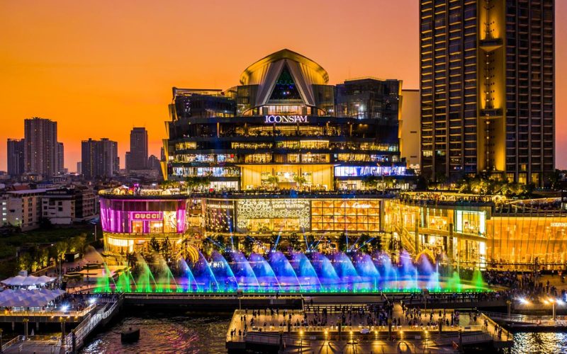 Le centre commercial Icon Siam illuminé au crépuscule, avec des jets d'eau colorés en mouvement et des boutiques luxueuses en arrière-plan