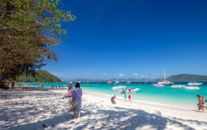 Plage de sable blanc en Thaïlande avec des eaux turquoise, des bateaux amarrés au large et des vacanciers profitant du soleil sous un ciel bleu éclatant