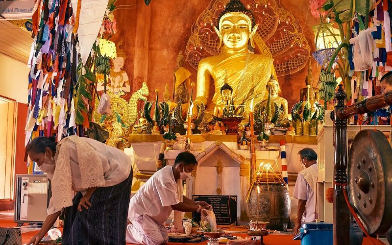 Des fidèles en tenue traditionnelle participent à une cérémonie bouddhiste dans un temple orné de statues dorées du Bouddha, d’offrandes et de décorations colorées
