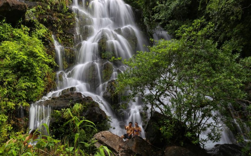 Cascade tropicale entourée de végétation luxuriante à Phong Nha, avec deux personnes assises sur un rocher au premier plan profitant de la vue et de la fraîcheur de l’eau