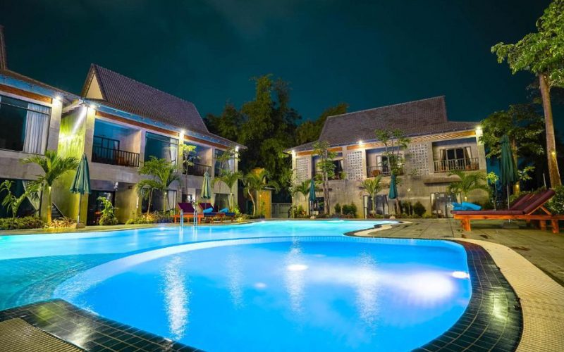 Photo d’un hôtel à Kampot de nuit, avec une grande piscine éclairée au centre, entourée de transats, de palmiers et de bâtiments à deux étages au style moderne et tropical