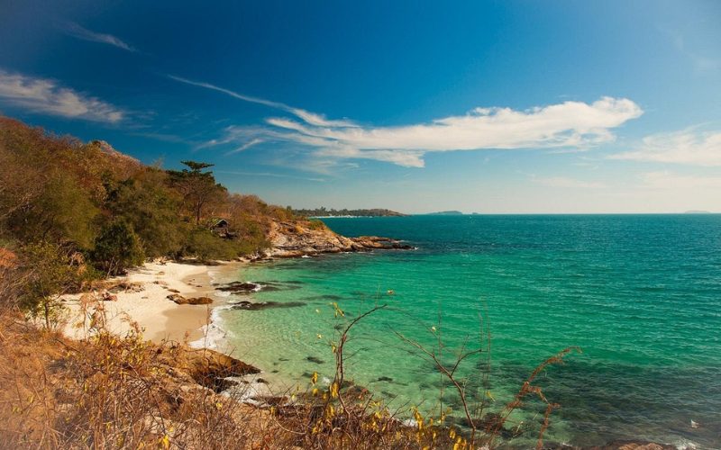 Plage isolée de sable blanc bordée de rochers et de végétation à Koh Samet, Thaïlande, avec une mer turquoise et un ciel bleu dégagé