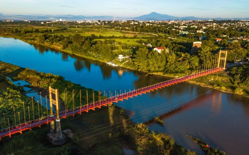 Vue aérienne d’un pont suspendu coloré traversant une rivière paisible à Kon Tum, entouré de paysages verdoyants et de la ville des Hauts Plateaux du Vietnam