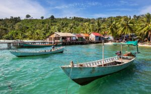 Barques traditionnelles amarrées sur une eau turquoise devant un petit village côtier aux maisons colorées, bordé de palmiers et entouré de végétation tropicale