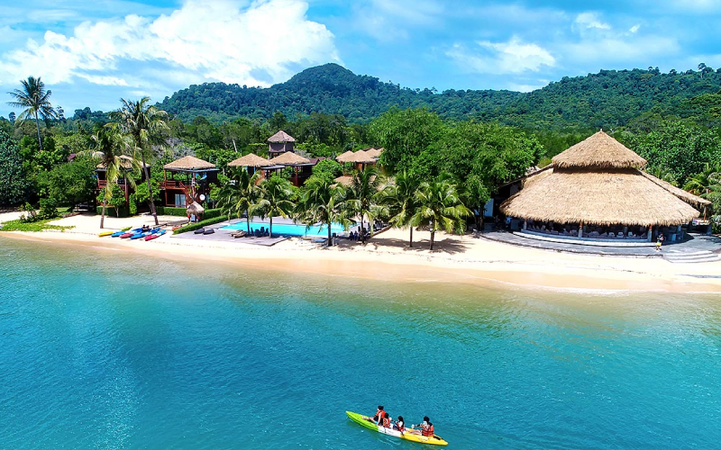 Resort en bord de mer avec plage de sable blanc, bungalows au toit de chaume, palmiers et kayak sur une eau turquoise, dans un décor tropical paisible