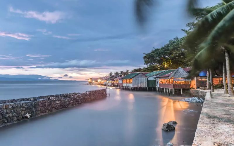 Vue en soirée de maisons sur pilotis éclairées le long d’un rivage paisible bordé de palmiers, avec un ciel bleu-gris au crépuscule et la mer calme reflétant les lumières dorées des bâtiments