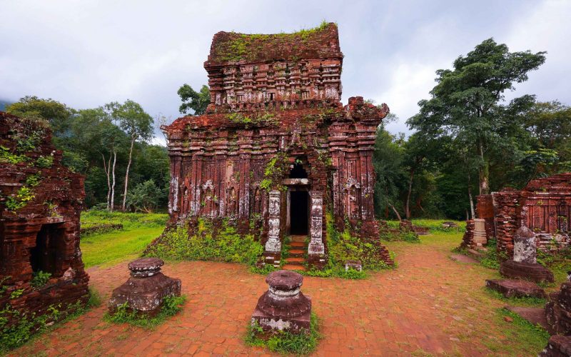 Ancien temple cham en brique rouge du sanctuaire de My Son, couvert de végétation, entouré d'arbres verdoyants sous un ciel nuageux
