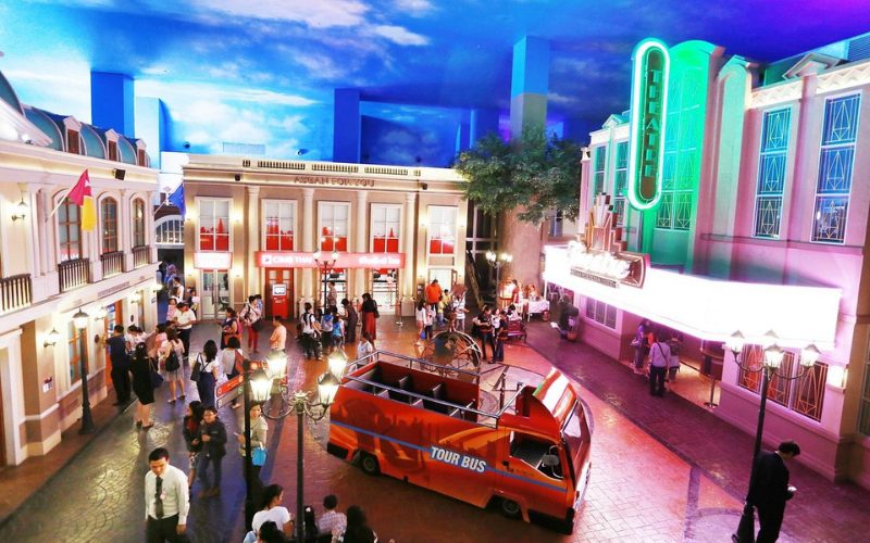 Vue intérieure de KidZania Bangkok, un parc éducatif où les enfants jouent à imiter différents métiers dans une ville miniature colorée, avec bâtiments, bus touristique et théâtre lumineux