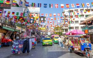 Rue animée de Khao San Road à Bangkok, décorée de drapeaux internationaux, avec des tuk-tuks colorés, des étals de vêtements et une atmosphère vibrante