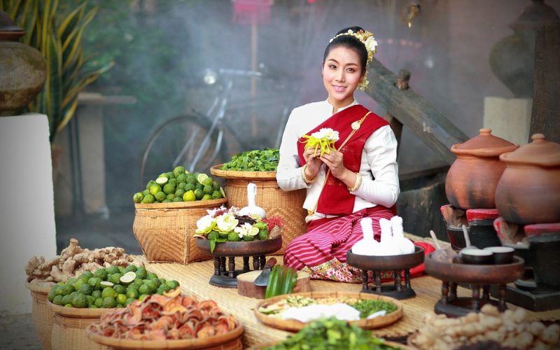 Femme vêtue d’un costume traditionnel Lanna préparant des herbes et fleurs pour un soin au spa à Chiang Mai, entourée de paniers remplis d’ingrédients naturels et d’épices