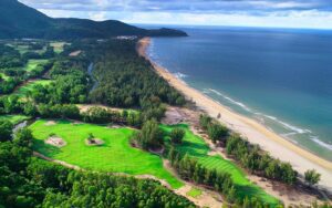 Vue aérienne spectaculaire du parcours de golf Laguna Lang Co au Vietnam, entouré de forêt tropicale et bordé par une longue plage de sable doré et l’océan