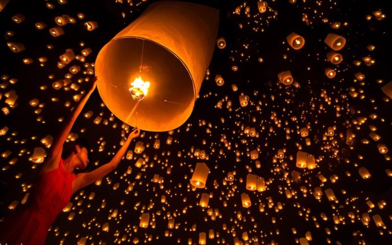 Une femme en robe rouge lâche une lanterne céleste illuminée dans le ciel nocturne, entourée de centaines d’autres lanternes flottant et créant une scène magique et chaleureuse