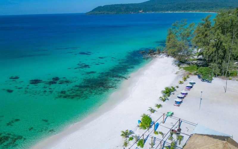 Plage de sable blanc immaculé bordée d’eaux turquoise à Koh Rong, avec des transats colorés, des palmiers et une mer calme s’étendant jusqu’à l’horizon