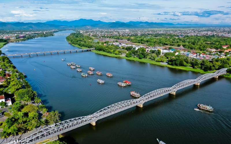 Vue aérienne de la rivière des Parfums à Hué, Vietnam, avec le pont Tràng Tiền emblématique et des bateaux traditionnels naviguant sur l'eau, entourés de verdure et de montagnes à l’horizon.