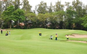 Des golfeurs et leurs caddies marchent sur un parcours de golf verdoyant, entouré d’arbres et de bunkers de sable, lors d’une journée nuageuse