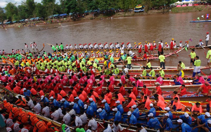 Vue aérienne de plusieurs équipes de rameurs vêtus de t-shirts de couleurs vives (orange, vert, rose, bleu, blanc, rouge) alignés dans de longues pirogues traditionnelles sur une rivière boueuse lors du Festival des pirogues