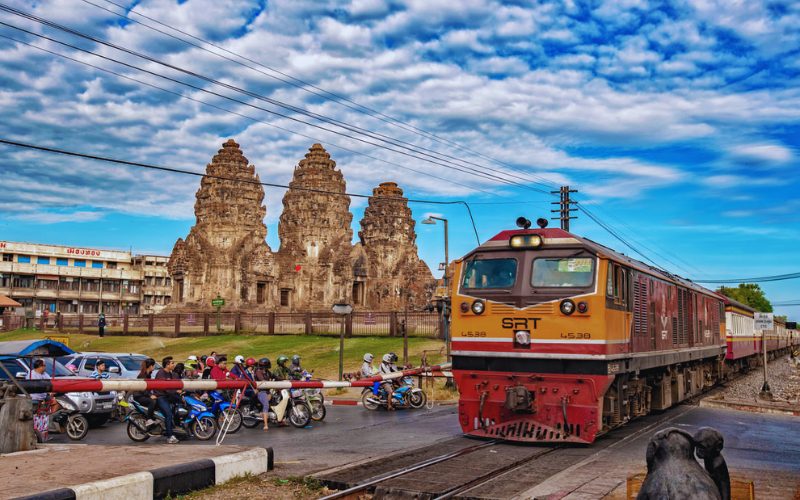 Vue emblématique de Lopburi avec le Phra Prang Sam Yot, un ancien temple khmer à trois tours, en arrière-plan, et un train SRT passant près d'un passage à niveau