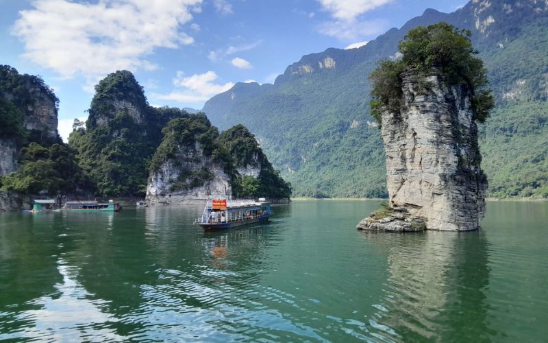 Paysage du lac Na Hang à Tuyen Quang, avec ses eaux vert émeraude entourées de montagnes boisées. On distingue une imposante falaise rocheuse solitaire émergeant du lac et un bateau de croisière naviguant paisiblement au milieu du décor naturel grandiose