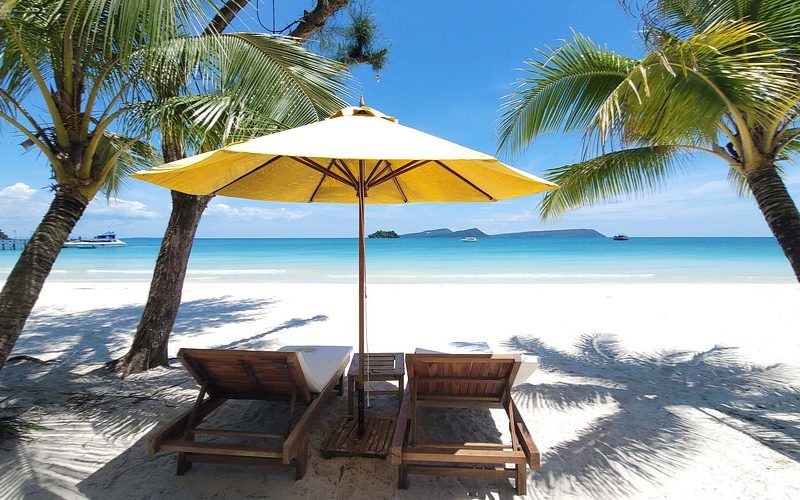 Deux transats en bois sous un parasol jaune, face à une plage de sable blanc bordée de palmiers et une mer turquoise à Koh Rong, Cambodg