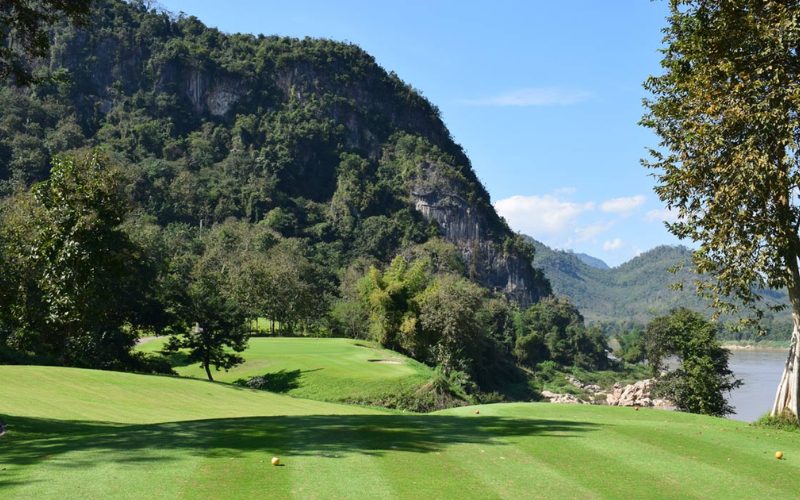 Parcours de golf verdoyant situé en bord de rivière, au pied d’une montagne couverte de forêt tropicale, sous un ciel bleu clair dans un décor naturel spectaculaire