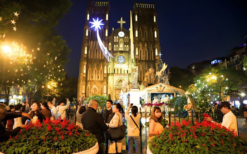 Foule rassemblée devant la cathédrale Saint-Joseph de Hanoï illuminée pour Noël, avec des décorations lumineuses, une crèche et une grande étoile scintillante dans le ciel nocturne