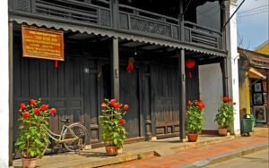 Façade d’une maison ancienne en bois dans le vieux quartier de Hoi An, avec un vélo et des pots de fleurs rouges alignés devant l’entrée