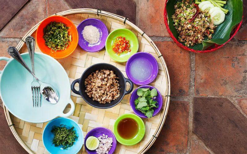 Plateau de cuisine traditionnelle laotienne présenté sur un panier en bambou, avec divers petits bols colorés contenant des herbes fraîches, épices, citron vert, sauce, viande hachée et légumes
