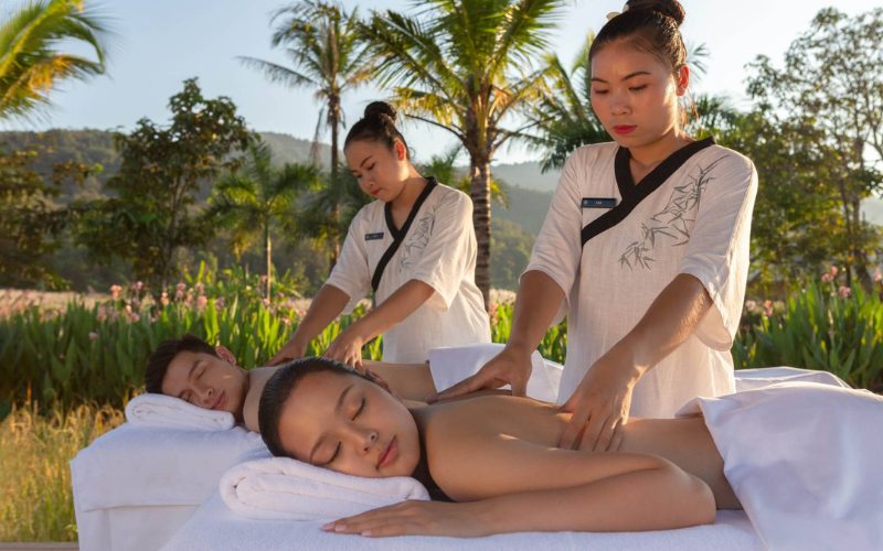 Deux personnes reçoivent un massage relaxant en plein air dans un cadre tropical luxuriant, entourées de palmiers et de collines verdoyantes, au coucher du soleil