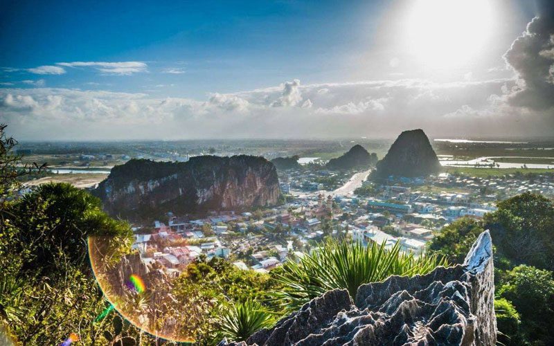 Vue panoramique sur Da Nang depuis un point de vue en hauteur, avec formations rocheuses, végétation au premier plan et soleil éclatant au-dessus de la vill