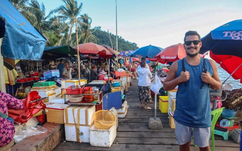 Touriste souriant dans le marché aux crabes de Kep, entouré d'étals colorés, de glacières et de parasols, avec une ambiance animée en bord de mer et des palmiers en arrière-plan
