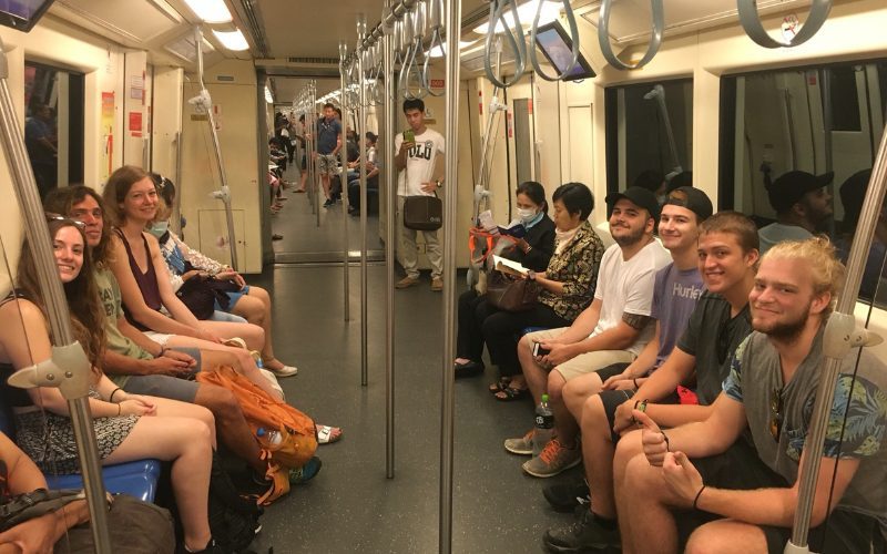 Intérieur d’une rame de métro à Bangkok avec des passagers assis sur les bancs latéraux, dont un groupe de jeunes touristes souriants et des habitants locaux