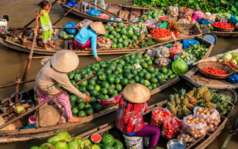 Un marché flottant animé sur une rivière du Vietnam, où des vendeurs dans des bateaux à rames traditionnels, coiffés de chapeaux coniques, vendent une grande variété de fruits et légumes frais, notamment de nombreuses pastèques et des ananas