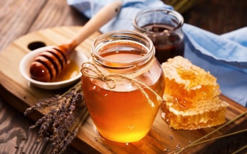 Pot de miel doré accompagné d’un rayon de miel frais, d’une cuillère en bois et de fleurs séchées, disposés sur une planche rustique en bois