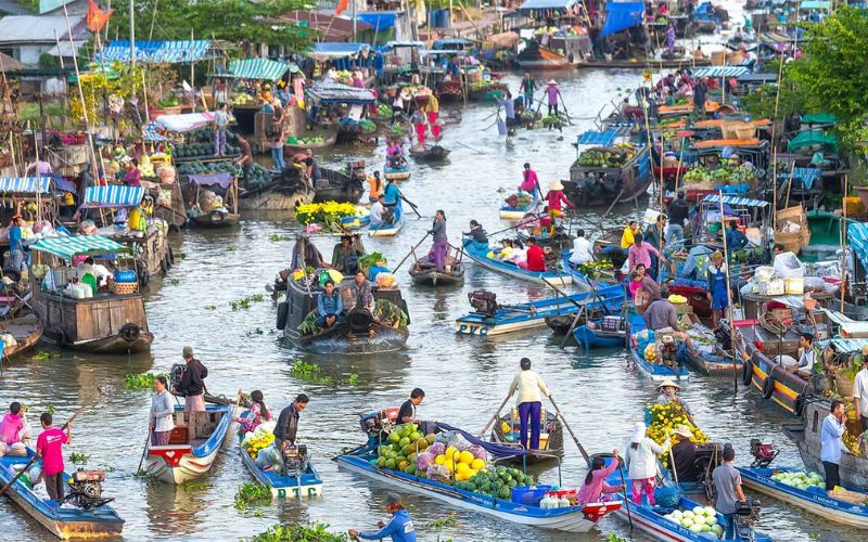 Vue animée d’un marché flottant dans le delta du Mékong, avec des dizaines de bateaux colorés chargés de fruits, légumes et fleurs, commerçants et acheteurs échangeant directement sur l’eau