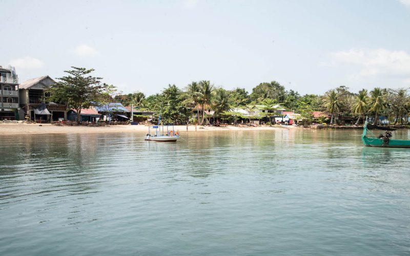 Vue sur le village côtier de M’Pai Bay à Koh Rong Sanloem, avec des maisons colorées, des palmiers et des bateaux traditionnels amarrés dans une eau calme.