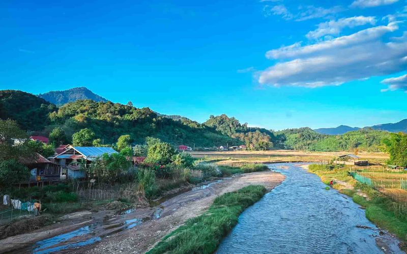Paysage paisible d’un village du Nord Laos, avec une petite rivière serpentant entre les rizières et les maisons traditionnelles sur pilotis, entouré de collines verdoyantes sous un ciel bleu clair