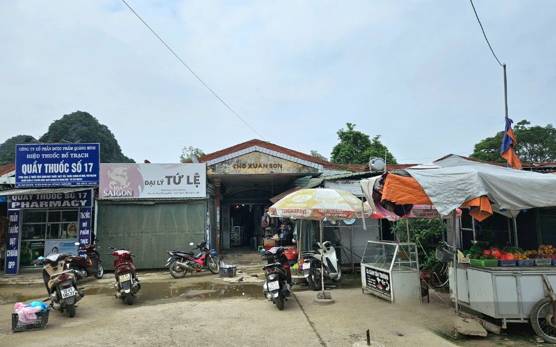 Entrée du marché Xuân Sơn à Phong Nha, avec des étals de rue, des scooters garés, une pharmacie locale et des vendeurs sous des parasols colorés
