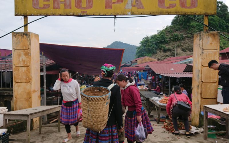 Scène animée au marché ethnique de Pà Cò, dans le nord du Vietnam, où des femmes Hmong en tenues traditionnelles colorées échangent et vendent des produits locaux sous les toits du marché