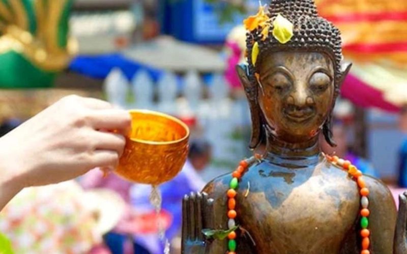 Main versant de l’eau parfumée sur une statue de Bouddha lors du Boun Pi Mai, avec des décorations colorées en arrière-plan pendant le festival lao