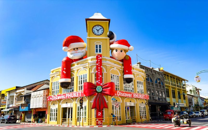 Bâtiment colonial jaune du centre historique de Phuket décoré pour Noël avec deux énormes personnages gonflables de Père Noël et d’un lutin, un grand ruban rouge et une banderole “Discover Phuket”, sous un ciel bleu éclatant