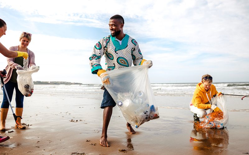 Groupe de bénévoles ramassant des déchets sur une plage, sacs en plastique remplis de détritus à la main, dans une ambiance conviviale et ensoleillée au bord de l’océan